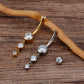 Two curved barbell belly button piercings with dangling clear rhinestone drops, one in gold-tone metal and one in silver-tone metal, displayed on a wooden surface with a small matching stud shown separately.