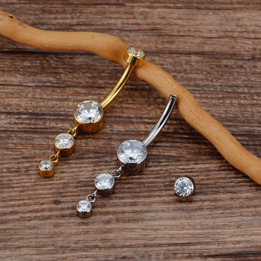 Two curved barbell belly button piercings with dangling clear rhinestone drops, one in gold-tone metal and one in silver-tone metal, displayed on a wooden surface with a small matching stud shown separately.