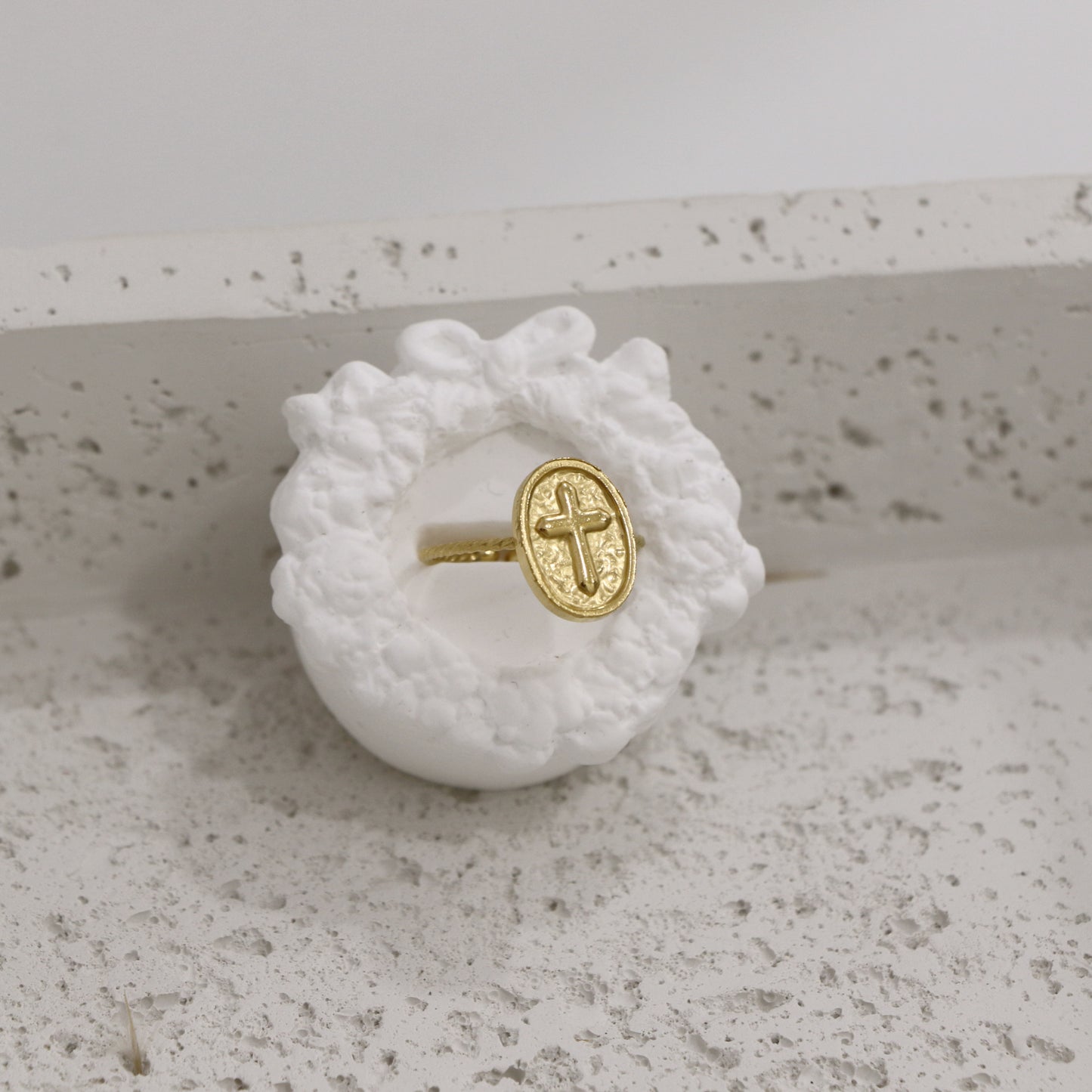 Gold ring with a cross design on a white textured stand against a light gray background