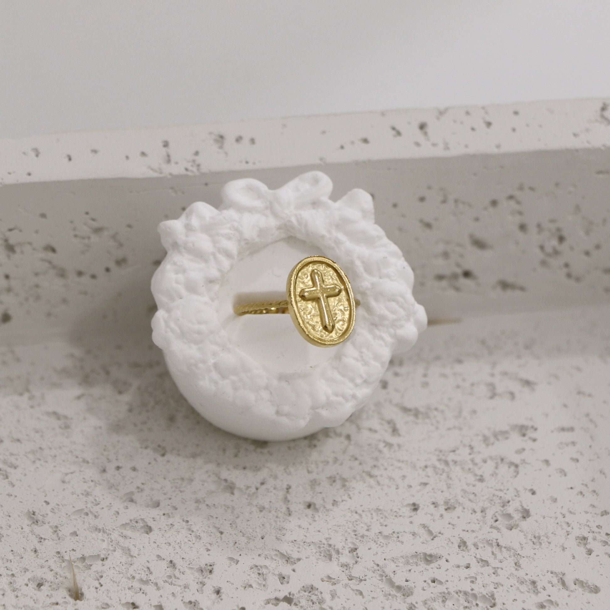 Gold ring with a cross design on a white textured stand against a light gray background