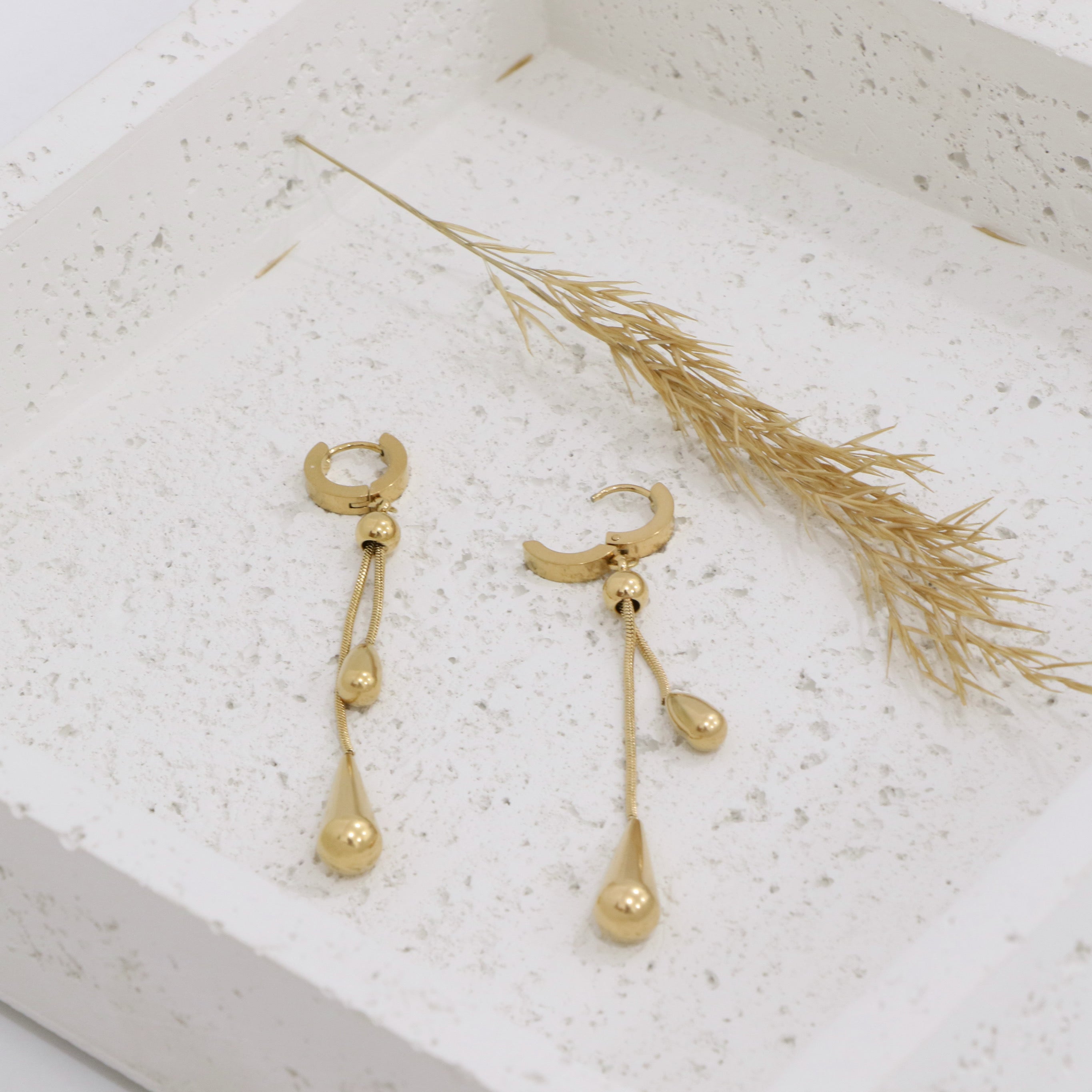 Gold earrings with dangling elements on a textured white surface with dried grass.