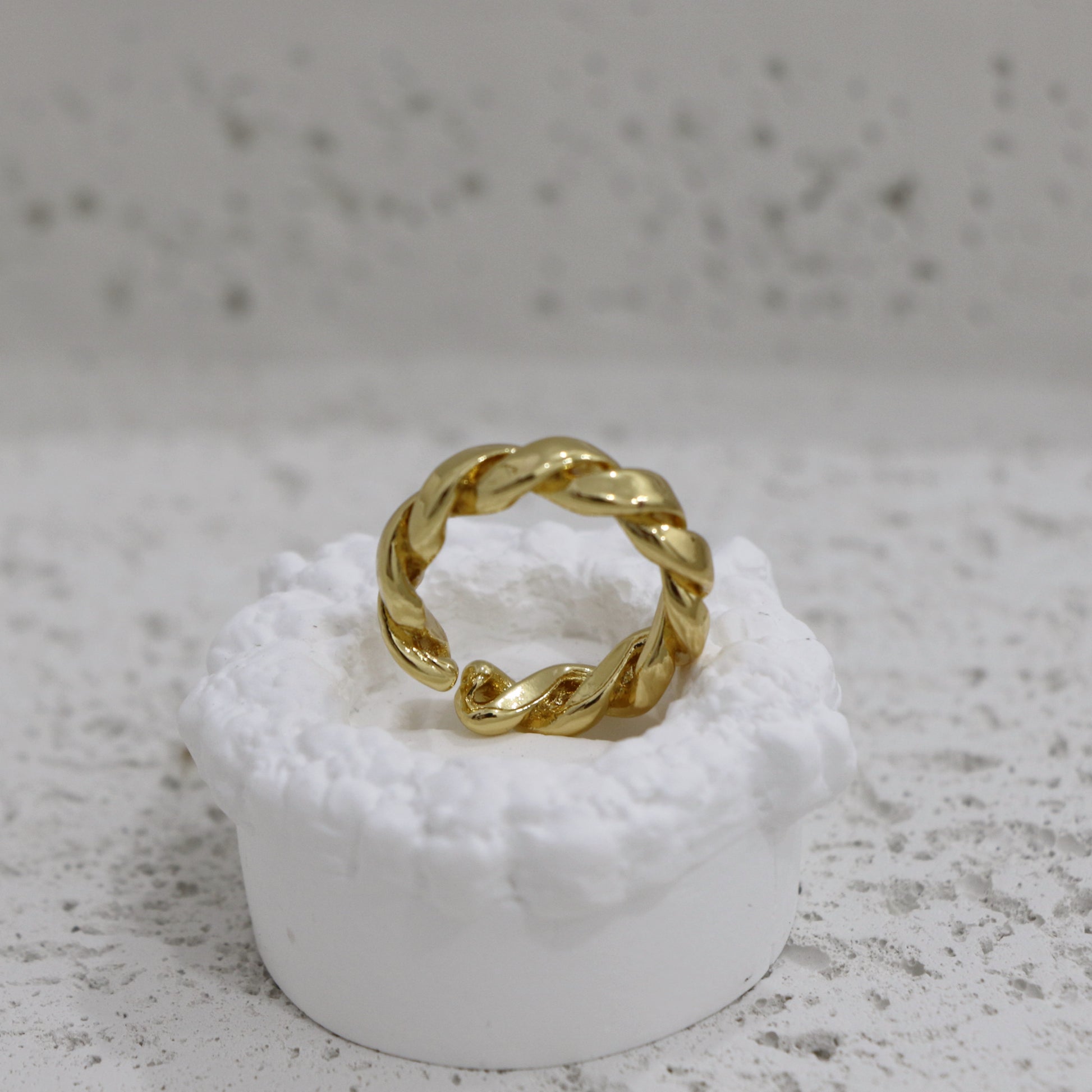 Gold braided ring on a white pedestal against a gray background