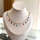 A gold chain necklace with small colorful teardrop charms displayed on a beige jewelry bust. The charms are in red, blue, green, black, and white.