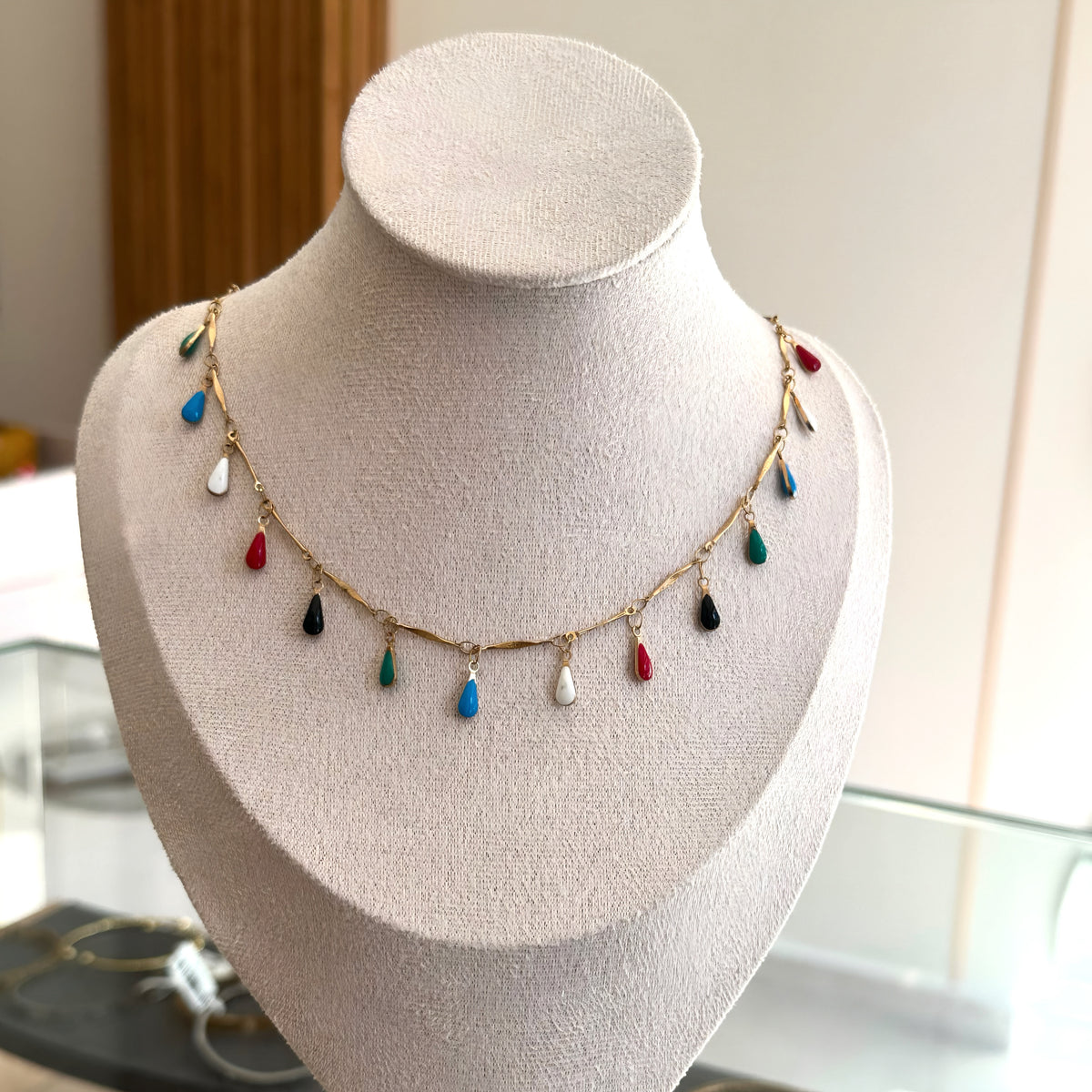 A gold chain necklace with small colorful teardrop charms displayed on a beige jewelry bust. The charms are in red, blue, green, black, and white.