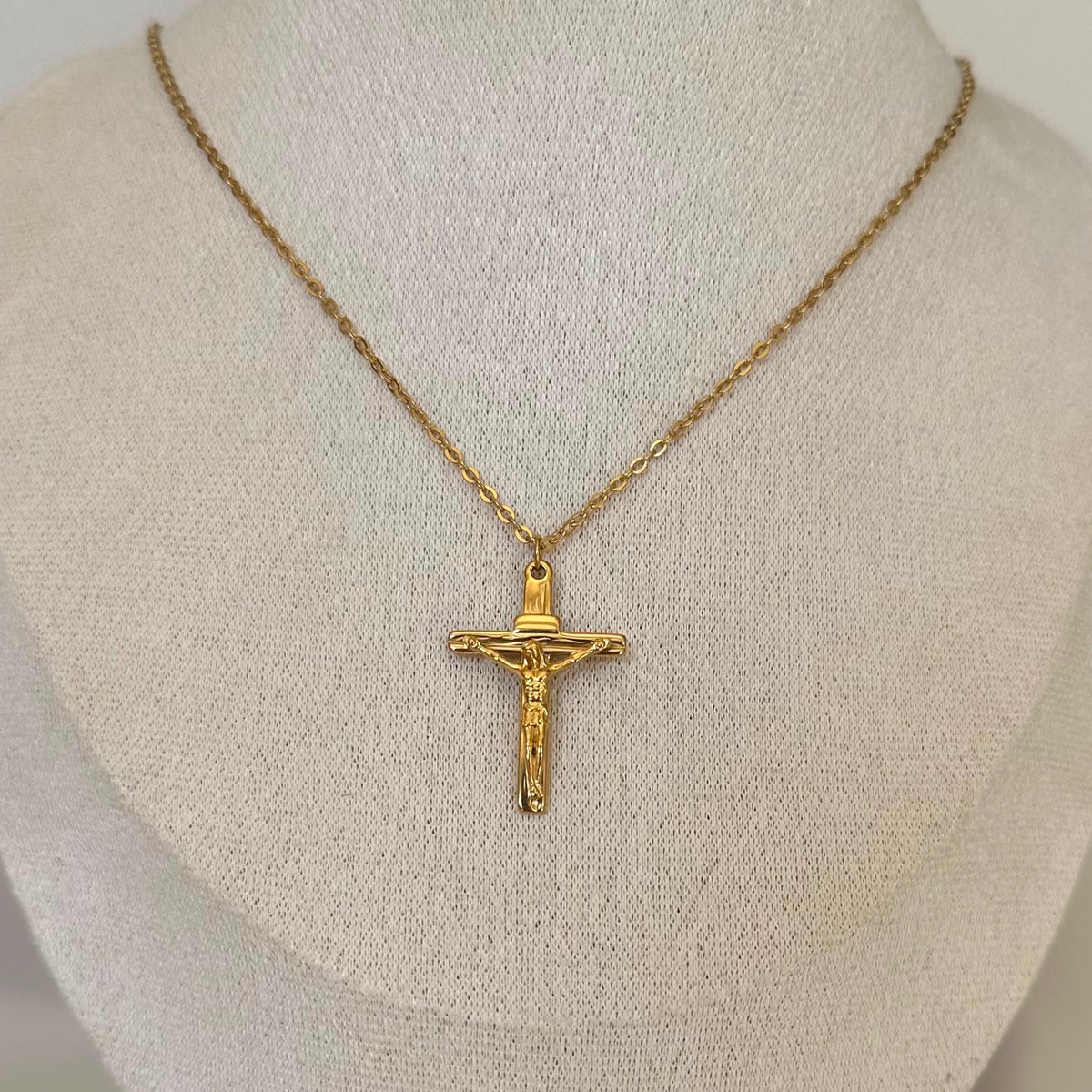 A gold crucifix pendant necklace on a textured cream fabric mannequin bust.