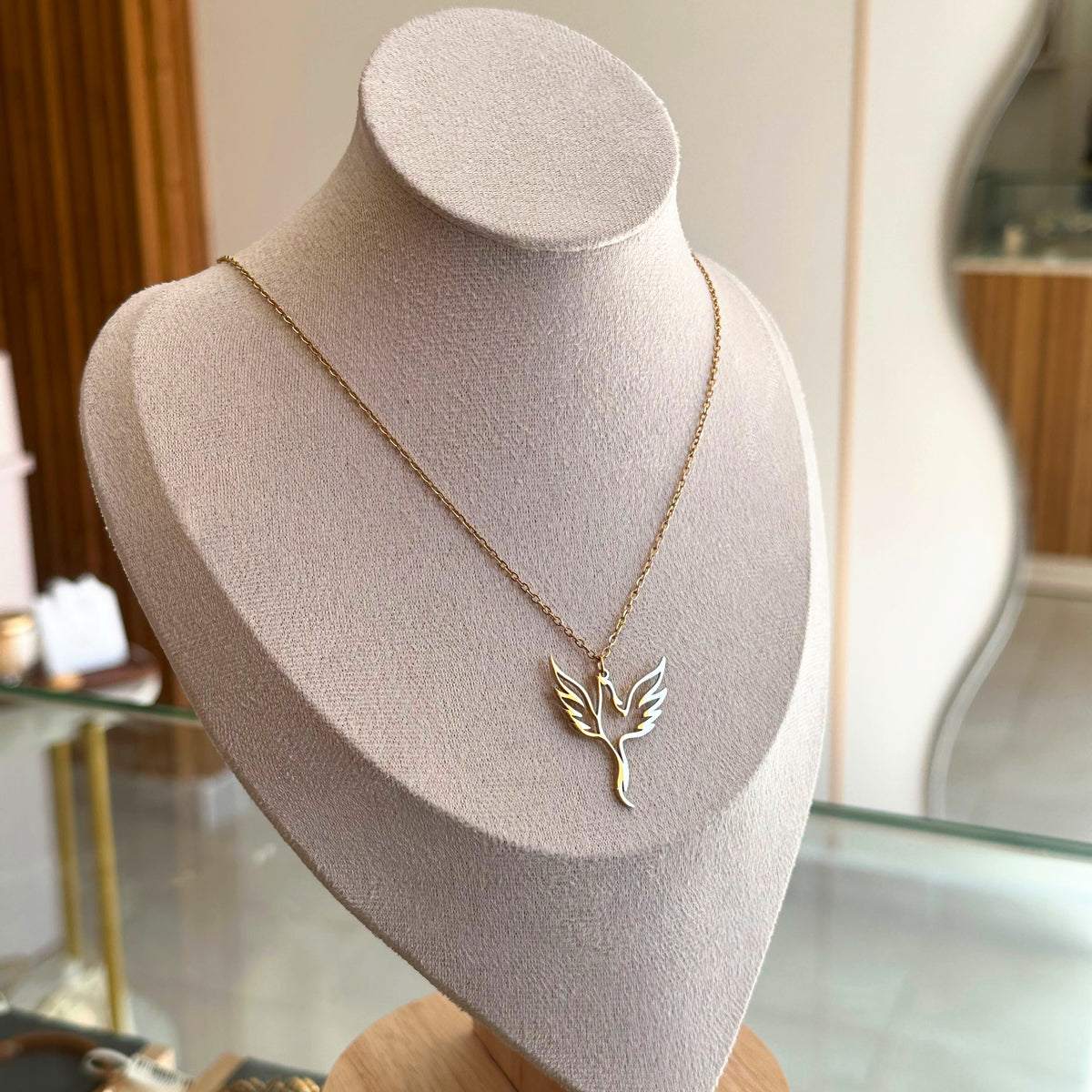 A delicate gold necklace with a phoenix shaped pendant displayed on a beige velvet jewelry bust in a boutique setting.