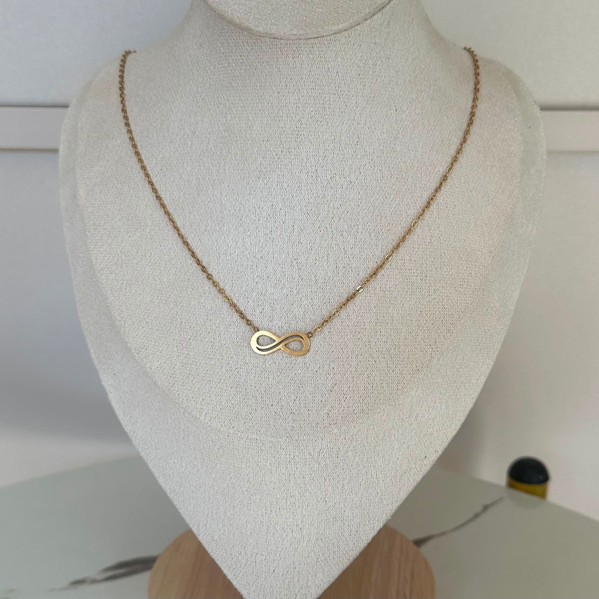 A delicate gold infinity necklace displayed on a textured cream-colored jewelry bust.